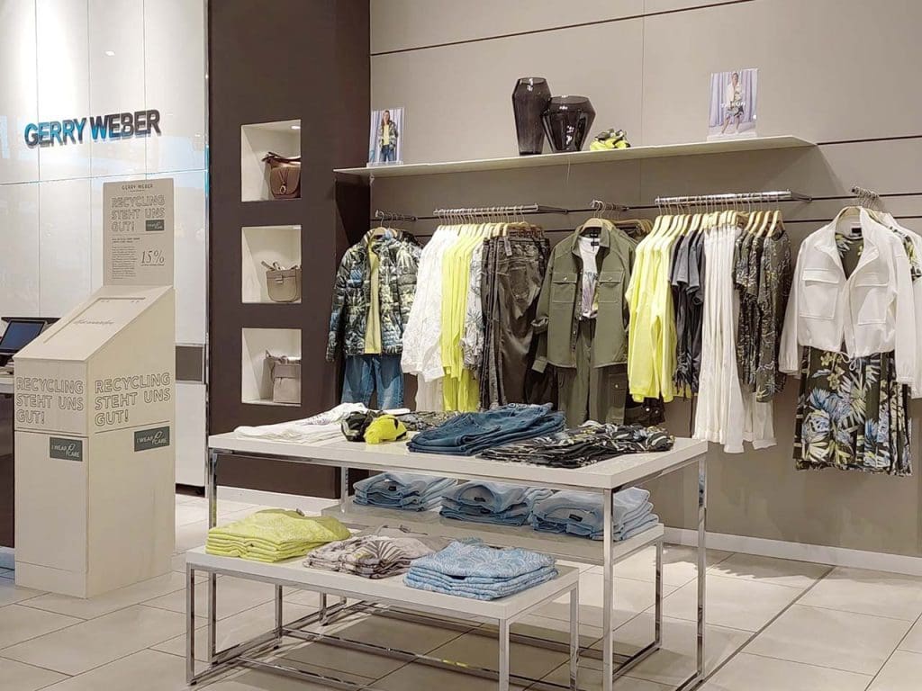 Bekleidung in den Farbtönen weiß, grün und gelb, hängend sowie liegend in einem Gerry Weber Store